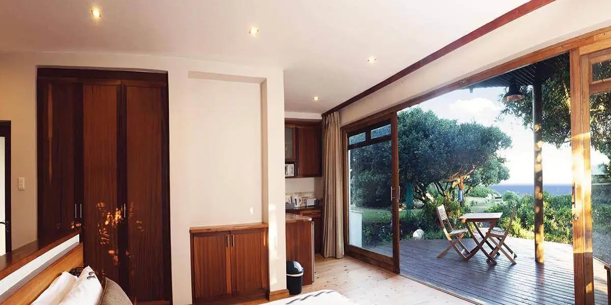 The Dolphin Luxury suite is a modern bedroom with a queen bed, wooden floors, with table and chairs, large windows with curtains, and an open bathroom area with a glass shower and natural light.