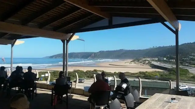 Wilderness fine dining while people relax at tables under a covered patio, enjoying Wilderness. The scene overlooks a sunny beach and ocean waves, with a coastal road and hills in the background beneath a clear blue sky.